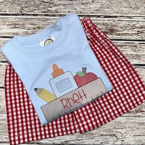 May include: A light blue t-shirt with a red, yellow, and green embroidered design of a pencil, glue bottle, and apple in a box. The name "Rhett" is embroidered below the box. The t-shirt is paired with red and white gingham shorts.