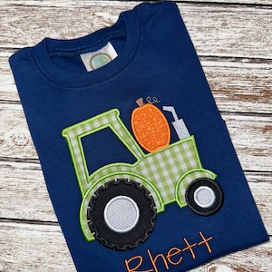 May include: A navy blue t-shirt with an embroidered green and white tractor carrying a pumpkin. The name "Rhett" is embroidered in orange below the tractor.