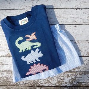 May include: A navy blue short-sleeved shirt with a green, blue, and red dinosaur applique. The shirt is paired with blue and white striped shorts.