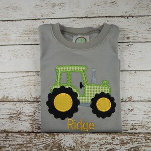 Puede incluir: Camiseta gris de manga corta con un tractor bordado en verde y amarillo y el nombre "Ridge" en amarillo.
