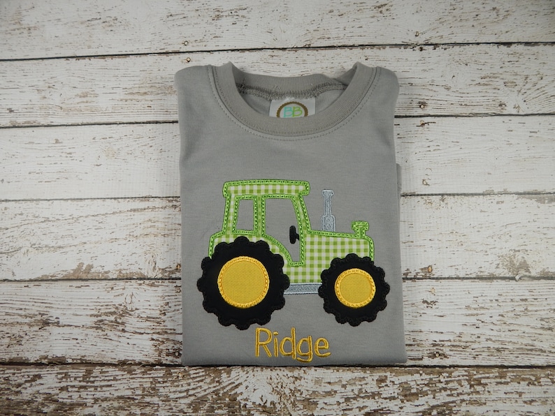 Puede incluir: Camiseta gris de manga corta con un tractor bordado en verde y amarillo y el nombre "Ridge" en amarillo.