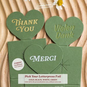 May include: Green heart-shaped cards with gold, white, and green foil lettering. The cards say "Thank You", "Vielen Dank", and "Merci". The cards are on a wooden surface with a daisy and the text "Pick Your Letterpress Foil".