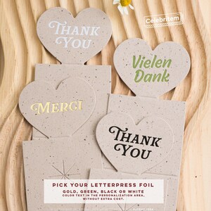 May include: Heart-shaped thank you cards with various text in white, gold, green, and black. The cards are on a textured beige background. The text "Pick your letterpress foil" is also visible.