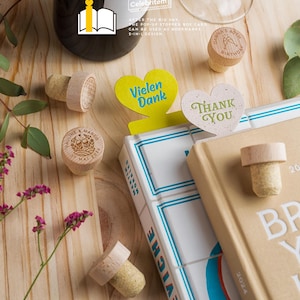May include: Wooden wine bottle stoppers with custom engravings and corks, alongside a book and decorative heart-shaped bookmarks. The image includes text that reads "Vielen Dank" and "Thank You". The product is a 2-in-1 design that can be used as bookmarks.