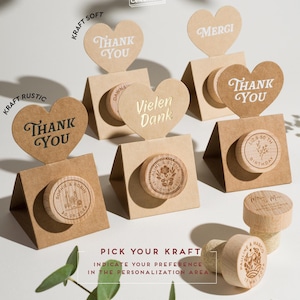 May include: A collection of heart-shaped kraft paper toppers with the words "Thank You" and "Merci" displayed above wooden wine stoppers. The stoppers are engraved with names, dates, and floral designs. The text "Pick Your Kraft" is also visible.