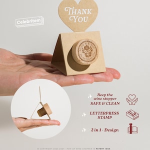 May include: A wooden wine stopper with a floral design and the names "Aline & Elena" and the date "26 October 2020" is displayed in a gift box with a heart-shaped "Thank You" tag. The stopper is held in a hand.