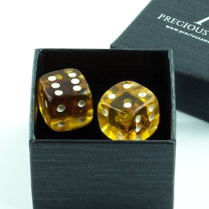 Op de afbeelding: Twee amber dobbelstenen met witte stippen in een zwarte doos met de tekst "Precious Amber" en het websiteadres "www.preciousamber.com".