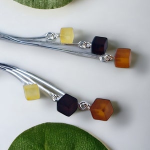 Può includere: Orecchini pendenti in argento con tre pietre di ambra in tonalità di giallo, marrone e nero. Le pietre hanno la forma di un cubo e sono attaccate agli orecchini con filo d'argento.