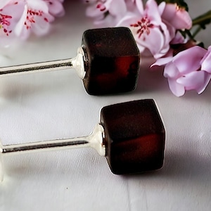 Könnte beinhalten: Ein Paar silberne Ohrstecker mit quadratischen Bernstein-Steinen. Die Steine sind in einem tiefen rotbraunen Farbton.