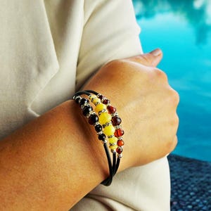 Puede incluir: Pulsera de múltiples hilos con cuentas de ámbar y plata. La pulsera tiene una banda de cuero negro y se usa en la muñeca. Las cuentas de ámbar son en tonos amarillos, naranjas y marrones. La pulsera es una joya.