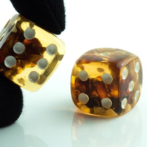 May include: Two amber dice with white dots. The dice are on a white surface. One die is being held by a black gloved hand.