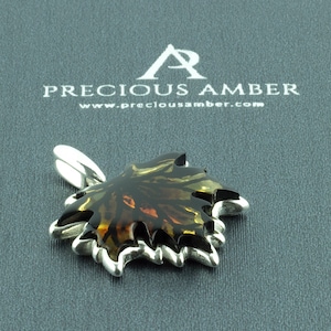 Puede incluir: Un colgante de hoja de arce de plata con una incrustación de ámbar marrón. El colgante está sobre un fondo gris con el texto "PRECIOUS AMBER" y "www.preciousamber.com".