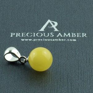 Può includere: Un ciondolo in ambra gialla con una balla d'argento. Il ciondolo è rotondo e liscio. Il ciondolo è su uno sfondo grigio con il testo "PRECIOUS AMBER" e "www.preciousamber.com".