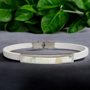 Könnte beinhalten: Weißes Lederarmband mit einer silbernen Leiste, die mit Perlmutt eingelegt ist.