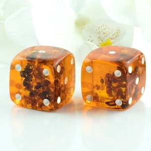 May include: Two amber dice with white dots. The dice are translucent and have small pieces of amber embedded in them.