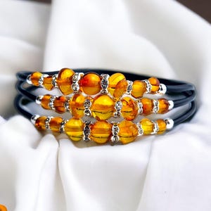 May include: A three-strand bracelet with amber-coloured beads and silver accents. The beads are interspersed with silver-toned spacers, and the bracelet is strung on black cords. The jewellery is displayed on white, silky fabric.