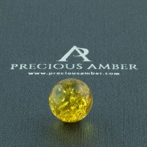 Può includere: Un perlino rotondo di ambra gialla con macchie dorate in tutto. Il perlino è su una superficie grigia con il testo "PRECIOUS AMBER www.preciousamber.com" sopra.