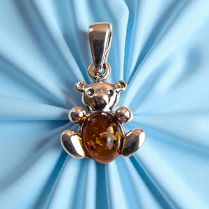 May include: Silver teddy bear pendant with a polished amber centre. The bear has rounded features and a loop for a chain. The pendant is set against a light blue background.