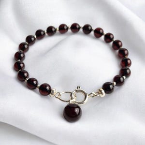Peut inclure: Un bracelet composé de perles rondes rouge foncé, reliées par des éléments métalliques argentés. Il comprend un charm rond assorti et une fermeture à fermoir. Les perles sont d'une couleur riche et profonde, présenté sur un tissu blanc et soyeux.