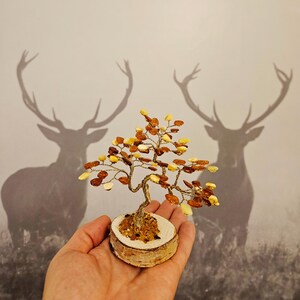 May include: A small amber tree sculpture on a birch wood base. The tree is made of wire and amber beads in shades of yellow and brown. The sculpture is held in a person's hand.