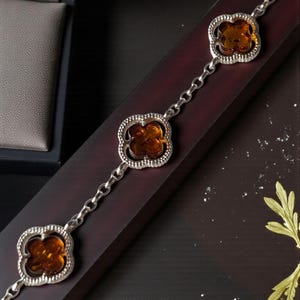 May include: Silver bracelet with three amber-coloured clover-shaped charms. The charms are set in a decorative silver frame and linked by a delicate chain. The bracelet is displayed on a dark wooden surface.