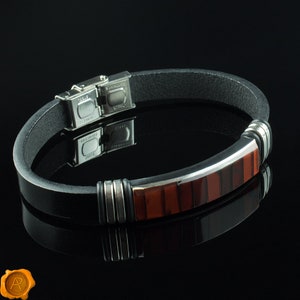 May include: A black leather bracelet with a silver and red inlay. The bracelet has a stainless steel clasp.