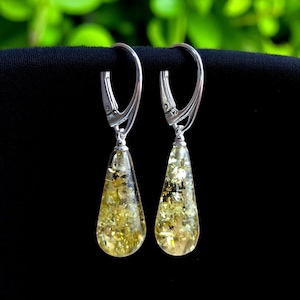 May include: A pair of dangle earrings with silver-colored leverback closures. The teardrop-shaped pendants are filled with golden-yellow amber and flecks. The earrings are displayed against a black background with green foliage in the background.