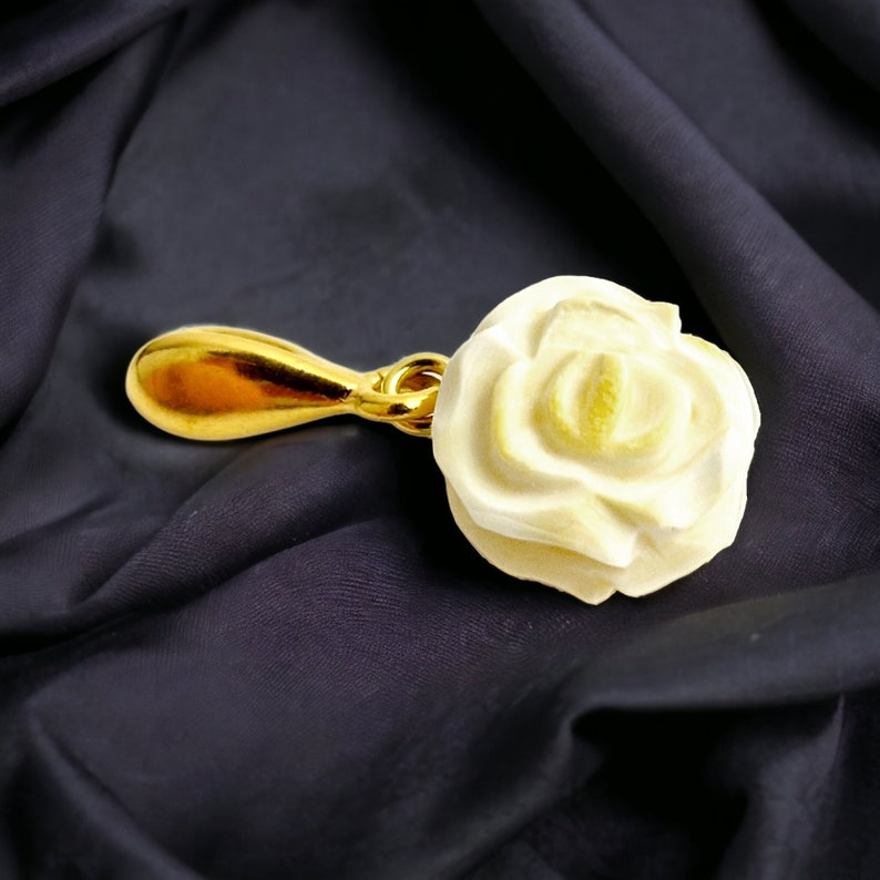 May include: A gold-colored pendant featuring a white rose-shaped charm. The pendant is on a black fabric background.