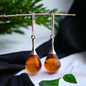 Op de afbeelding: Zilveren oorhangers met druppelvormige barnsteen hangers. De barnsteen heeft een warme, goudbruine kleur en is gefacetteerd om het licht te vangen. De oorbellen hangen aan zilveren haken.