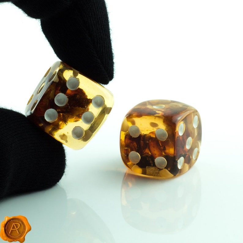 May include: Two amber dice with white dots. The dice are on a white surface. One die is being held by a black gloved hand.