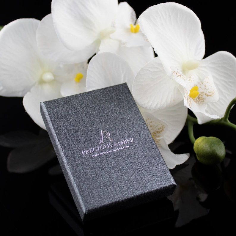 Puede incluir: Una caja rectangular gris con el texto "Precious Amber" y la direcci&oacute;n del sitio web "www.preciousamber.com" impresa en la parte superior. La caja est&aacute; sobre una superficie negra con flores blancas en el fondo.