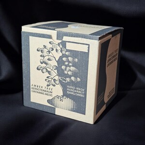 May include: A cardboard box with a black and white illustration of a tree. The box has text that reads "Amber Tree, Bernsteinbaum, Gintarinis Medis, Hand-Made, Handarbeit, Ranku Darbo".
