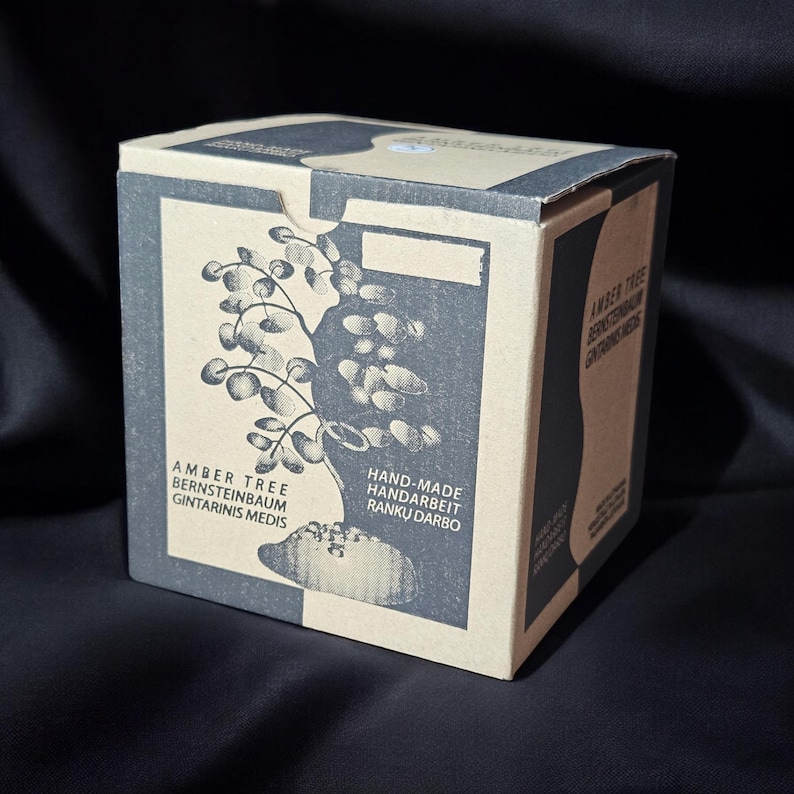 May include: A cardboard box with a black and white illustration of a tree. The box has text that reads "Amber Tree, Bernsteinbaum, Gintarinis Medis, Hand-Made, Handarbeit, Ranku Darbo".