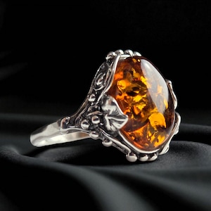 Könnte beinhalten: Ein Silberring mit einem großen, ovalen Bernstein. Der Bernstein hat eine warme, durchscheinende orange Farbe mit inneren Mustern. Der Ring hat ein verziertes, floral inspiriertes Design um den Stein und am Band.