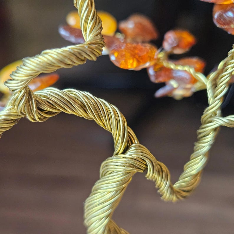 May include: A gold wire tree sculpture with amber gemstones. The wire is twisted and knotted to create a realistic tree trunk and branches. The amber gemstones are scattered throughout the branches, adding a touch of color and sparkle.