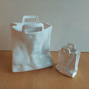 May include: Two white ceramic planters shaped like paper bags. The larger planter is taller and wider than the smaller planter.