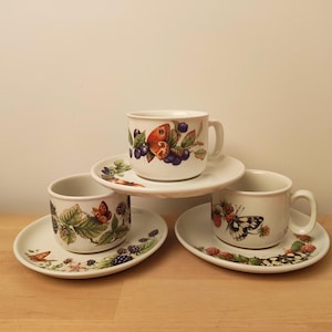 Peut inclure: Trois tasses à thé et soucoupes en céramique blanche avec des motifs de papillons et de baies. Chaque tasse présente un arrangement différent de papillons et de baies. Les soucoupes ont un motif de bordure assorti. Les tasses sont empilées.