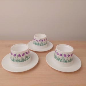 Peut inclure: Trois coquetiers en céramique blanche avec soucoupes assorties. Chaque tasse présente une bande de fleurs violettes et de gazon vert. Les tasses sont disposées sur une surface en bois clair.
