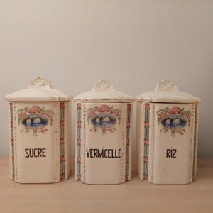May include: Three off-white ceramic canisters with lids. Each canister has a floral and swan design, with the words "SUCRE", "VERMICELLE", and "RIZ" in black text. The canisters are hexagonal.