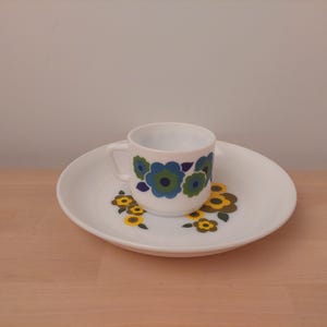 Könnte beinhalten: Ein weißes Keramik-Tassen- und Untertassen-Set. Die Tasse und die Untertasse weisen ein Blumenmuster in Blau-, Grün- und Gelbtönen auf. Das Set steht auf einer hellbraunen Holzoberfläche.