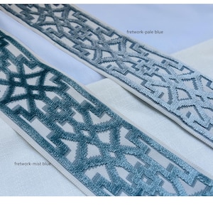 May include: Two samples of trim fabric with a geometric pattern. The trim on the top is a pale blue color and the trim on the bottom is a darker blue color. The text "fretwork-pale blue" is written above the top trim and the text "fretwork-mist blue" is written below the bottom trim.