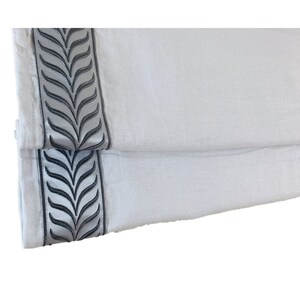 May include: A pair of white linen roman shades with a gray embroidered leaf design along the bottom edge.