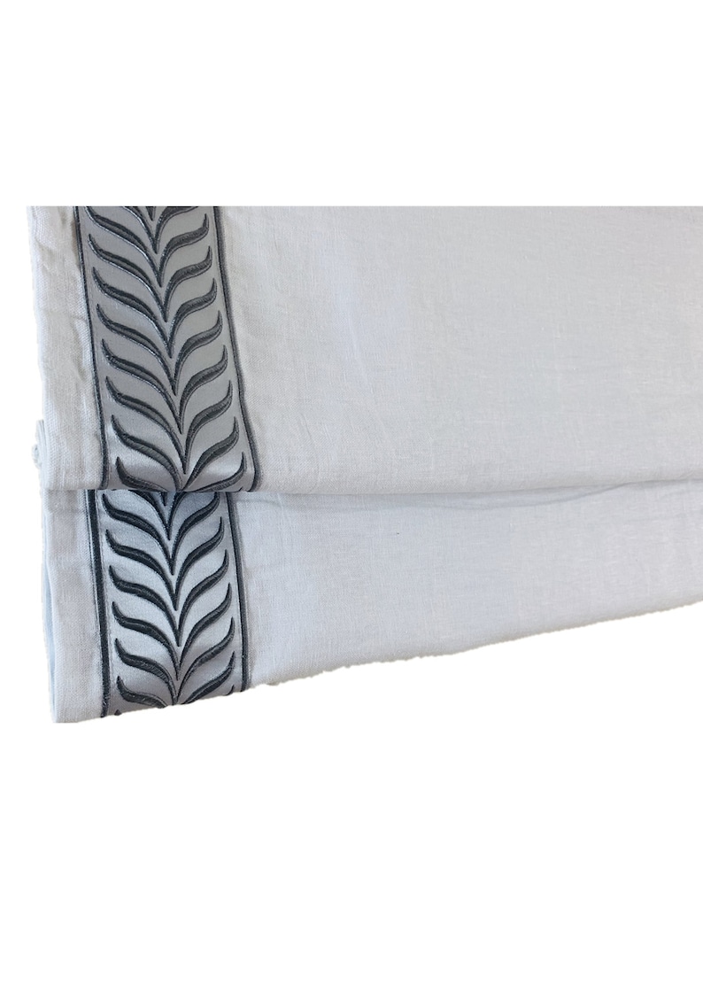 May include: A pair of white linen roman shades with a gray embroidered leaf design along the bottom edge.