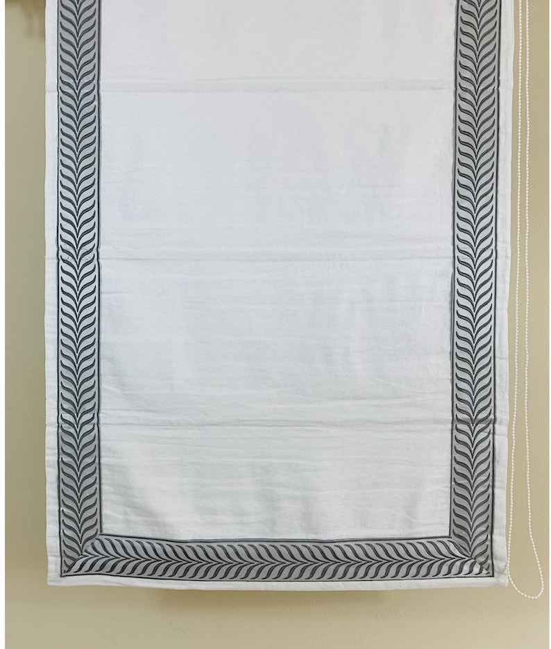 May include: White roman shade with a gray leaf pattern border. The shade is pulled up to the top of the window.