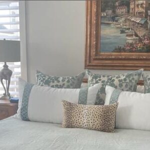 May include: A white bed with a light blue patterned comforter and pillows. The bed has a wooden headboard and is decorated with a variety of pillows, including a patterned pillow with a floral design, a solid white pillow, and a patterned pillow with a geometric design. There is a lamp on a wooden nightstand next to the bed.