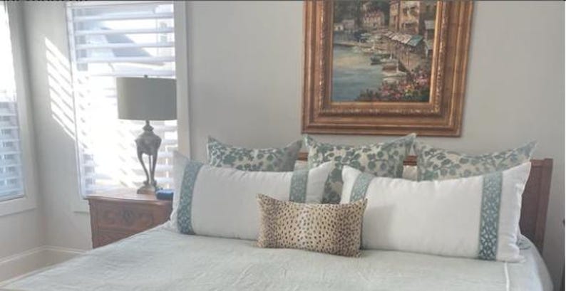 May include: A white bed with a light blue patterned comforter and pillows. The bed has a wooden headboard and is decorated with a variety of pillows, including a patterned pillow with a floral design, a solid white pillow, and a patterned pillow with a geometric design. There is a lamp on a wooden nightstand next to the bed.