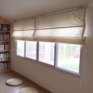 May include: A set of white roman shades with a light texture hang in a window with a view of trees and greenery. Two round woven mats are on the floor below the window.