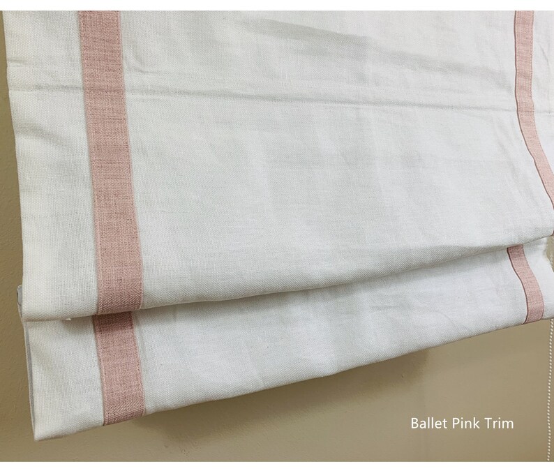 May include: White roman shade with a pink trim. The trim is a woven fabric with a subtle texture. The shade is folded in half, showing the trim on both sides. The text "Ballet Pink Trim" is visible at the bottom of the image.