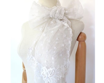 May include: A white lace scarf with a delicate floral pattern. The scarf is tied in a bow around the neck.
