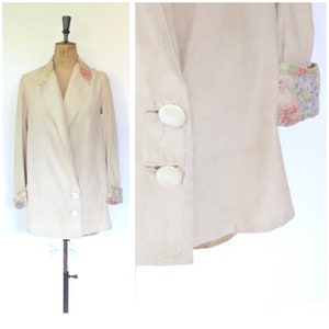 May include: A cream linen jacket with floral trim on the collar and cuffs. The jacket has a shawl collar, a single-breasted closure, and two large white buttons.